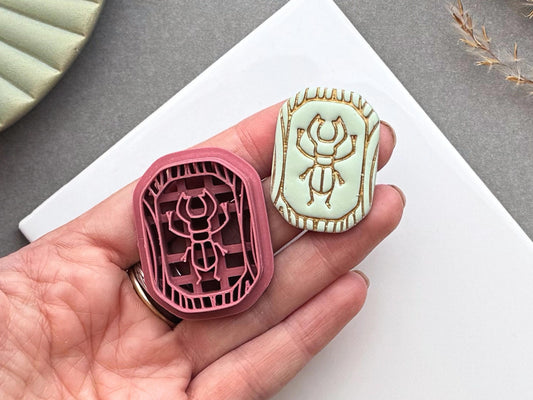 Scarab beetle polymer clay cutter and matching finished polymer clay embellishment held in a hand for scale, featuring a detailed scarab beetle within a decorative Egyptian-inspired border.