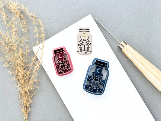 Three specimen jar-style scarab beetle clay cutters in different colours, featuring a crescent moon, detailed scarab beetle and small star details. A finished cream polymer clay scarab cut-out is shown alongside the pink and blue cutters. Gothic and celestial-inspired design.