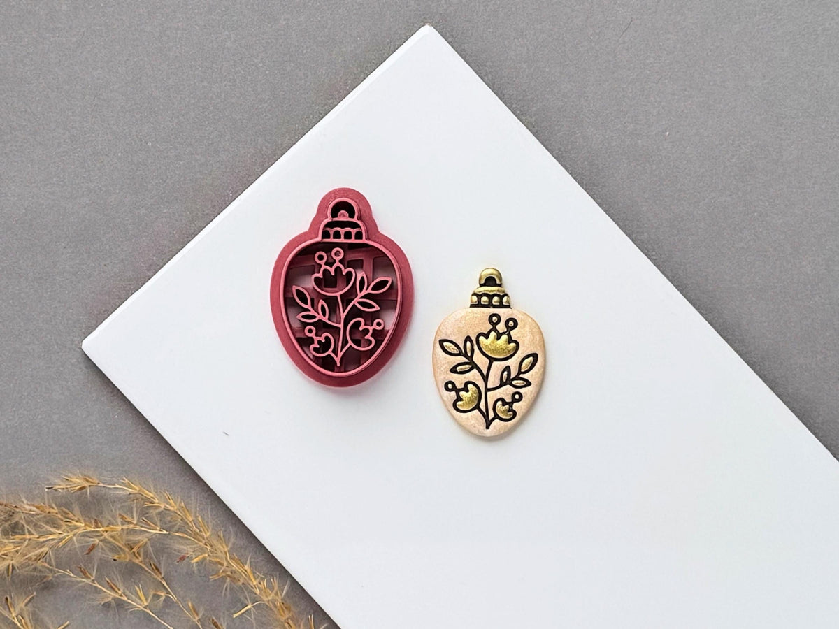 Floral Christmas bauble polymer clay cutter displayed beside a matching finished clay embellishment featuring a decorative botanical design with flowers, leaves and curling stems.