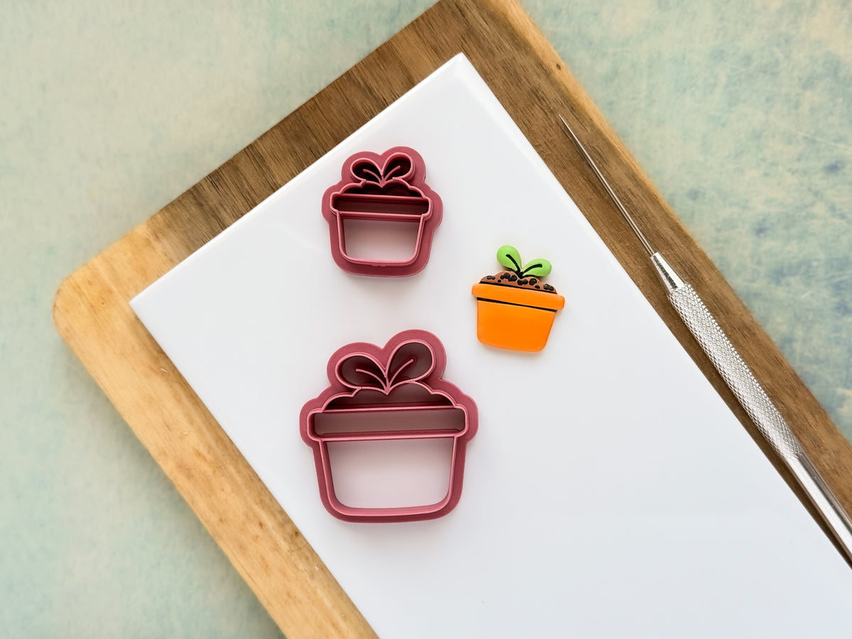 Flower pot shaped clay cutters with soil and seedling details in large and small sizes. Next to the cutters is a polymer clay sample cut out painted in orange, brown and green.