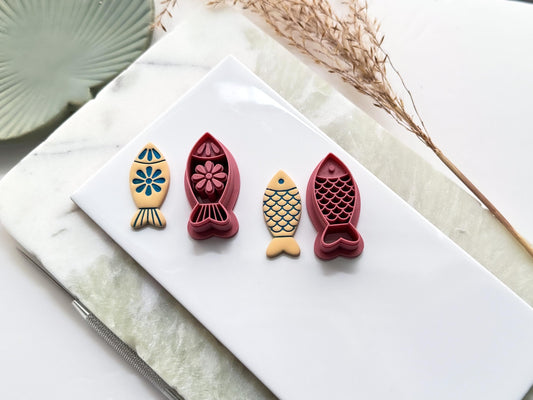 Dark pink coloured simple fish shaped clay cutters, one cutter has a folk style floral pattern, and the other has a scales pattern. Next to them are polymer clay sample cut outs made with the cutters and painted. All are on a white tile with decorative items.