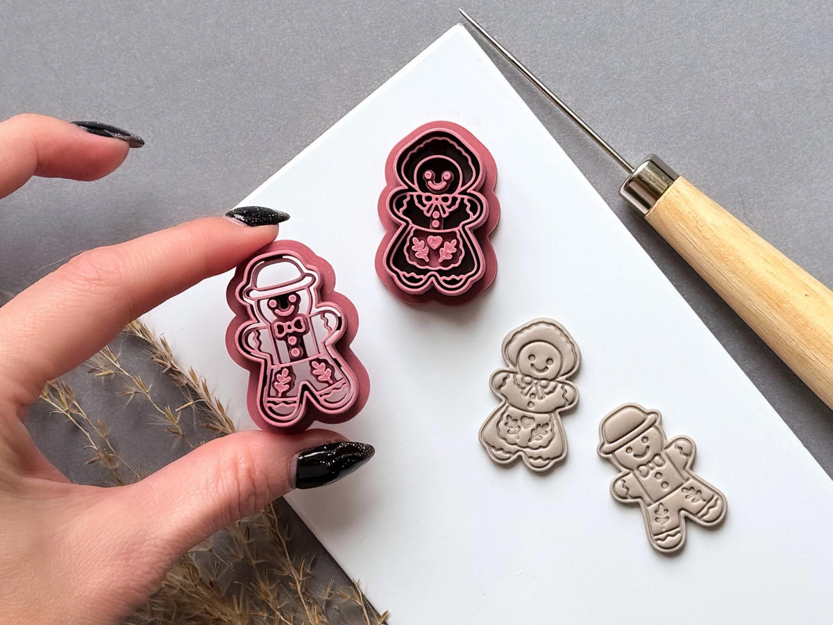 Vintage-inspired gingerbread man and gingerbread girl polymer clay cutters featuring folk-art floral motifs, decorative icing details, hats, bow ties and smiling faces. One cutter is held in a hand to showcase the detailed stamp design, with matching finished clay embellishments displayed on a white tile.