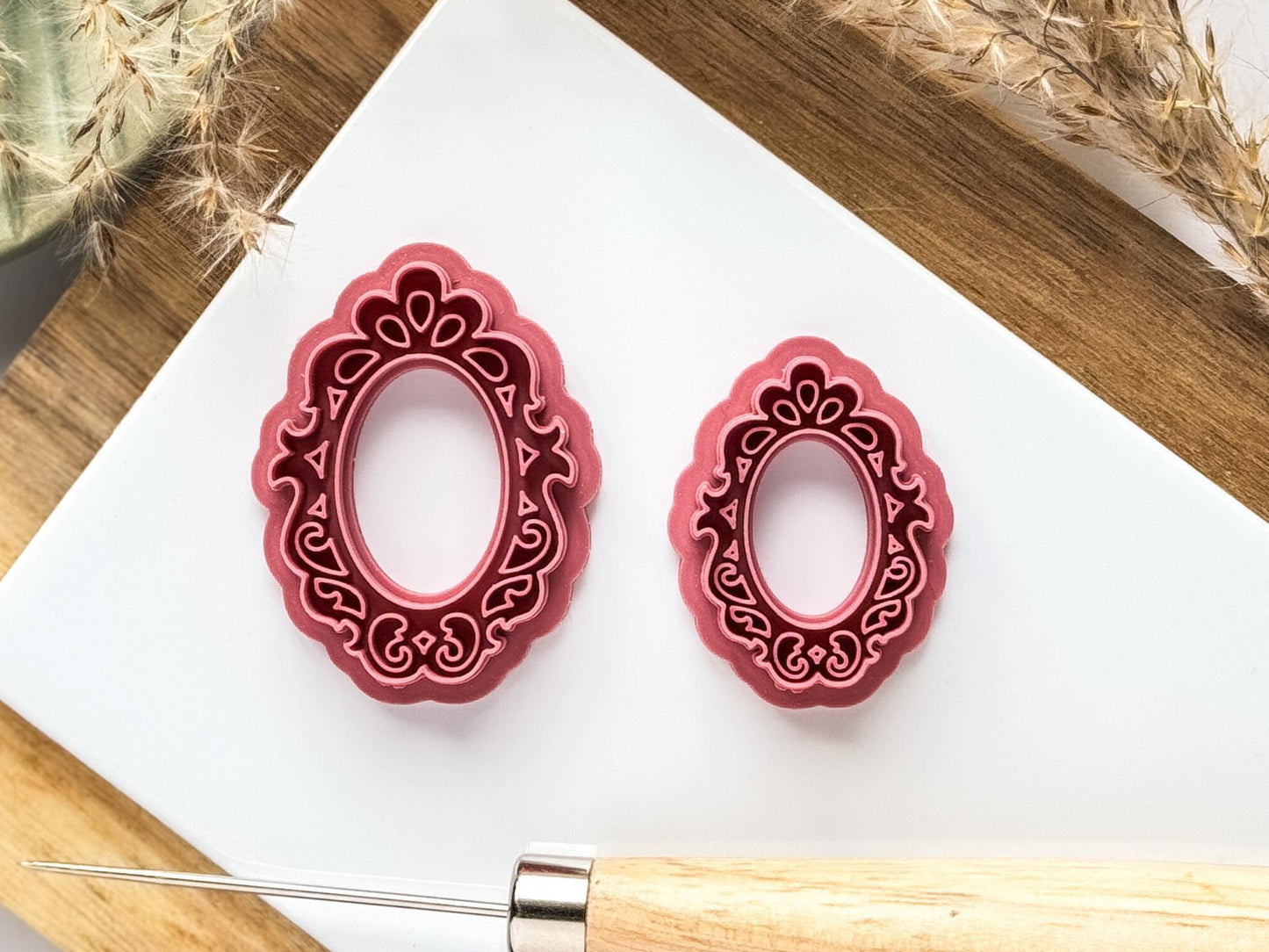 Small and large pink filigree mirror clay cutters on a white clay baking tile surrounded by ornamentsand clay tools