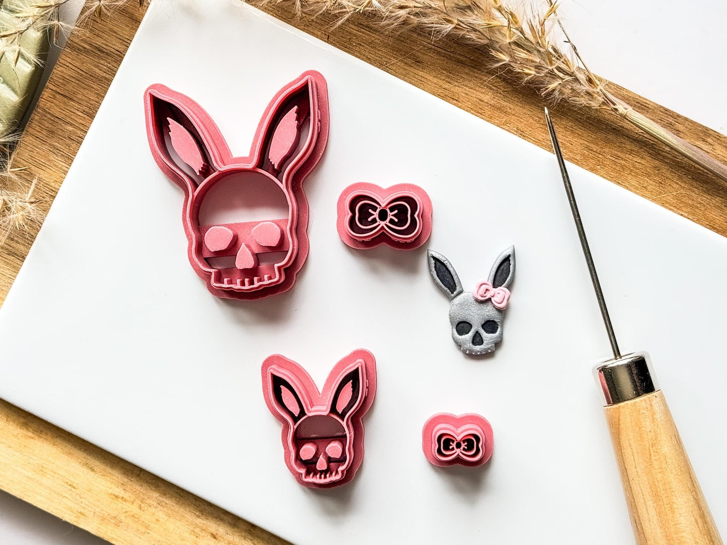 Skull clay cutter in pink with rabbit ears and separate bow shaped nano cutter with cut out clay sample on a white clay baking tile with a tool.