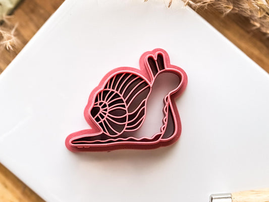 Close up image of a single snail shaped 3D printed polymer clay cutter in dark pink on a white tile