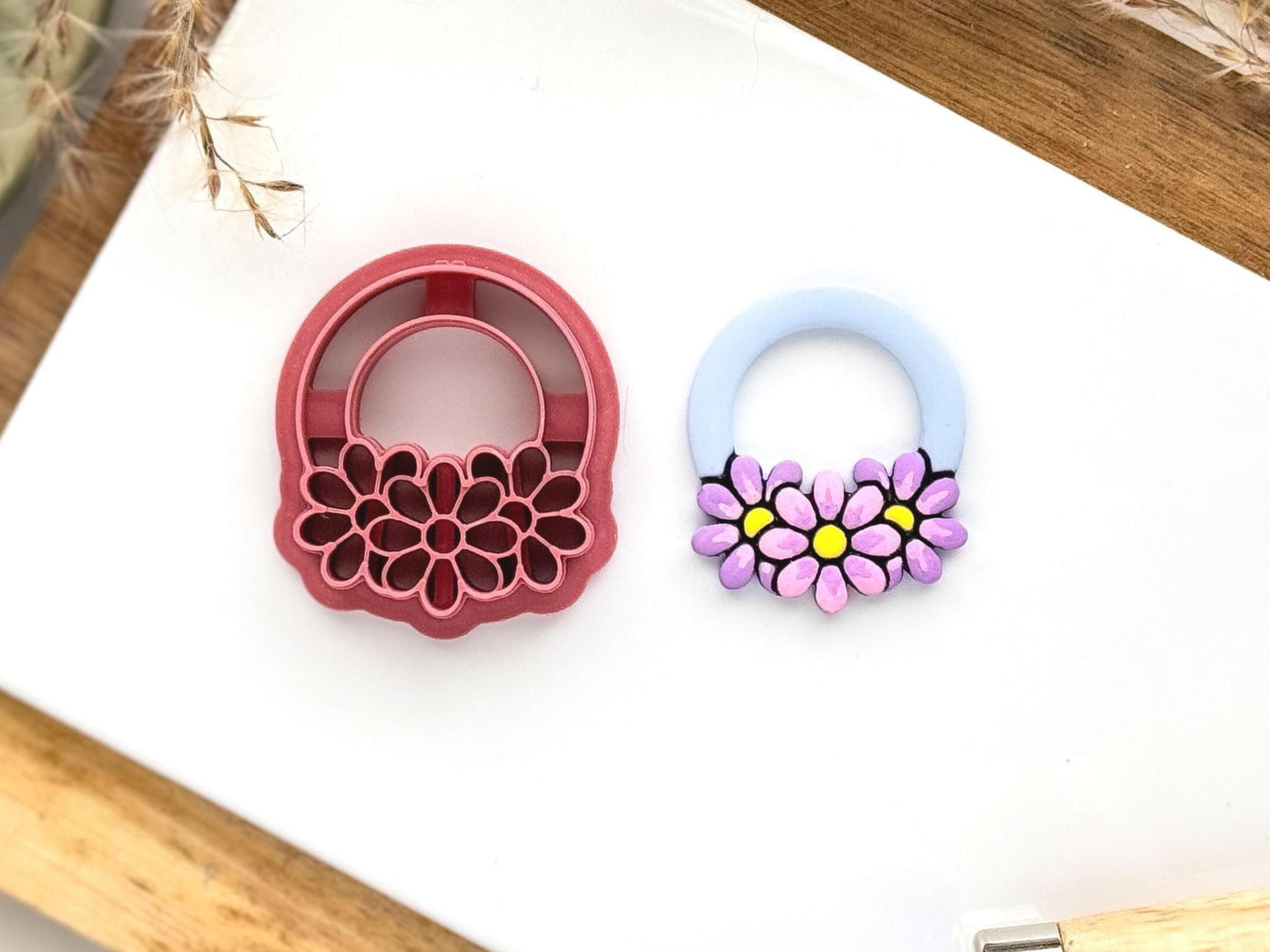 Triple daisy flower and ring polymer clay cutter in dark pink on a white tile background. Next to the cutter is a cut out polymer clay sample in baby blue, pink and purple.