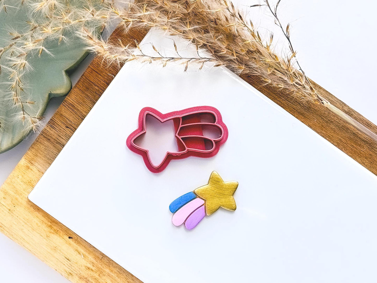Shooting star shaped clay cutter with five point star and 3 trails separated by imprint lines. Next to the cutter is a gold sample clay cut out with with blue, pink and purple coloured star dust trails
