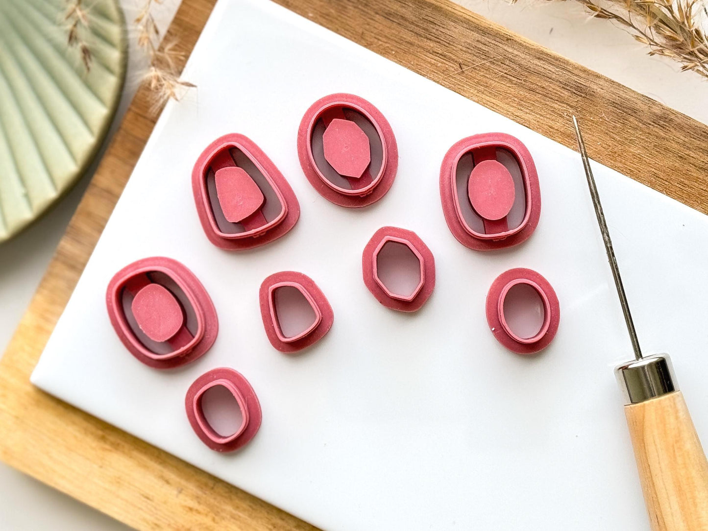 Geometric shapes inlay clay cutter set in dark pink on a white ceramic baking tile and wooden chopping board. Sourrounded by a green trinket dish and clay tool