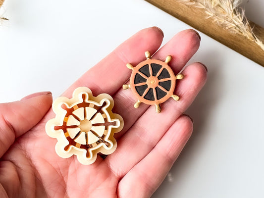 Ships helm clay cutter with debossedsections, raised wooden spokes and golden handles being held in hand for scale