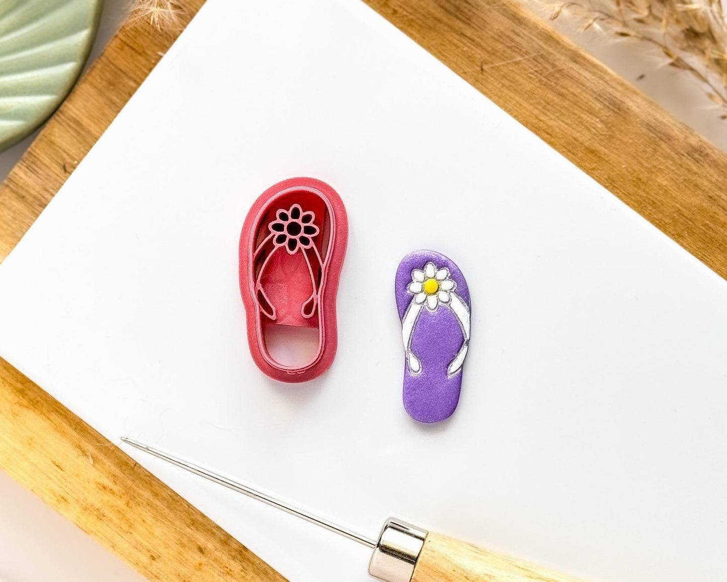 Polymer clay cutter in the shape of a summer flip flop with straps and flower detail next to a purple flipflop cut out clay sample with painted details.