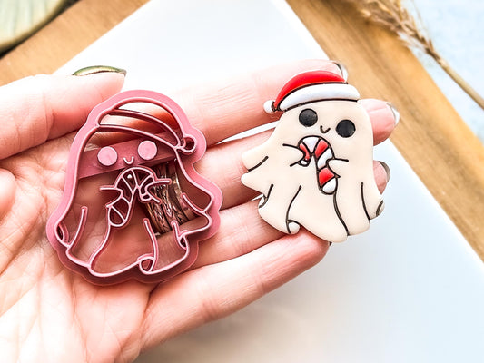 A pink plastic clay cutter shaped like a ghost wearing a Santa hat, holding a candy cane. Beside it is a finished ghost-shaped clay earring with a Santa hat and candy cane. The ghost is white with black eyes.