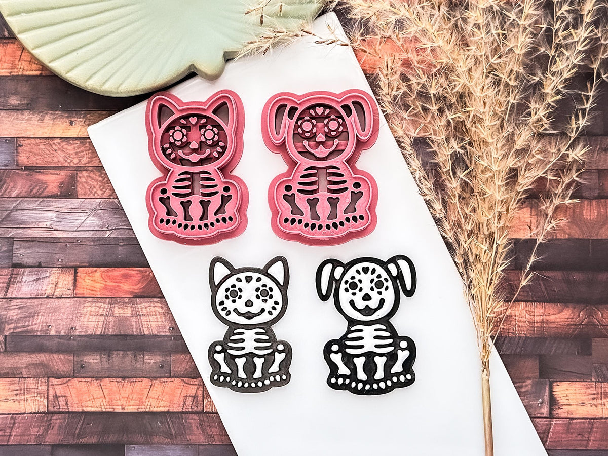 Pink sugar skull style dog and cat cutters with embossing bone details, sample polymer clay cut outs in black and white next to them on a white tile