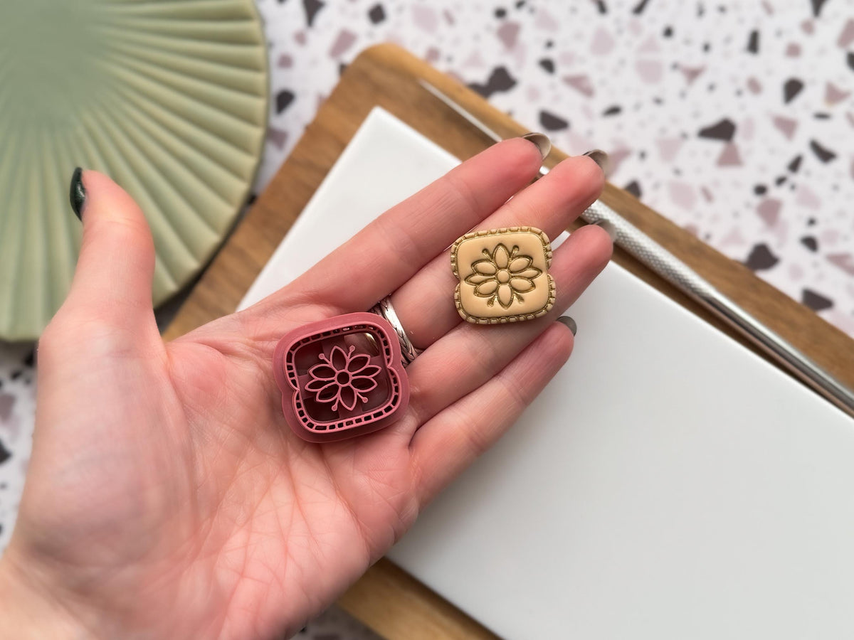 this image contains a small pink polymer clay cutter in a rectangular shape with rope pattern outer edge detail and a folk art style flower in the middle being held in a hand for scale