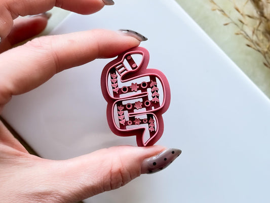 Pink coloured snake-shaped polymer clay cutter with intricate floral stamp details in the style of boho or folk art. The cutter is being held between two fingers
