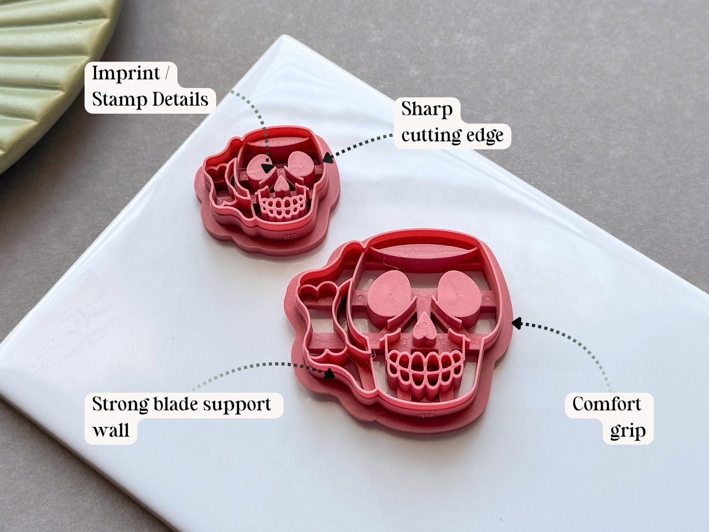 Side view of a skull shaped mug polymer clay cutter in pink PLA, highlighting the sharp cutting edge, reinforced support wall, embossed stamp details and comfortable grip handle. Displayed on a white tile with decorative dried grasses.