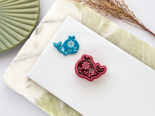 Red floral whale-shaped polymer clay cutter and matching blue clay cut-out displayed on a white ceramic tile over a marble-style background, showcasing intricate floral stamp details for jewellery and craft making.