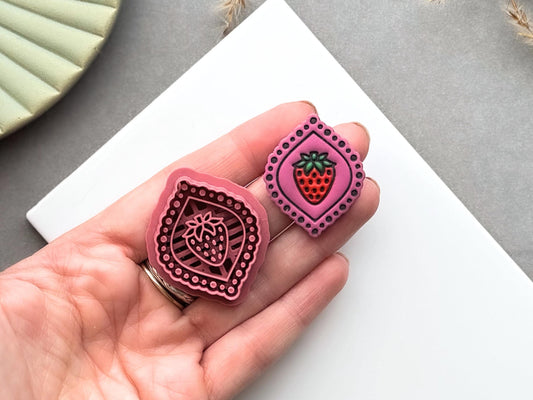 Strawberry frame polymer clay cutter and matching finished polymer clay embellishment held in a hand for scale, featuring a strawberry inside a decorative beaded frame, perfect for summer fruit, berry and botanical clay designs.
