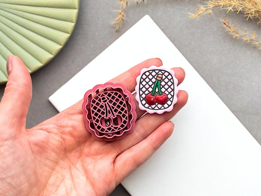 Cherry polymer clay cutter displayed beside a matching finished polymer clay embellishment featuring two cherries on a decorative lattice background within a scalloped frame.