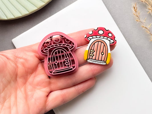 Mushroom cottage polymer clay cutter and matching finished clay embellishment held in a hand for scale, featuring a whimsical red-and-white spotted mushroom with a tiny arched door, perfect for cottagecore, woodland, fairycore and whimsical clay projects.