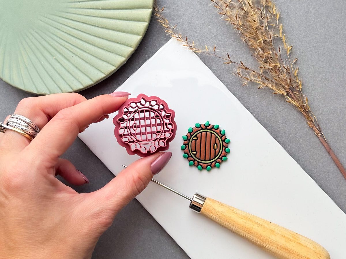 Pink round fairy door clay cutter held in hand beside a green and brown fairy door clay piece, featuring a circular wooden door design with stone and foliage details, photographed on a white clay work surface with dried grasses and a green plate. Cottagecore, woodland, autumn, fairy garden and seasonal clay craft design.