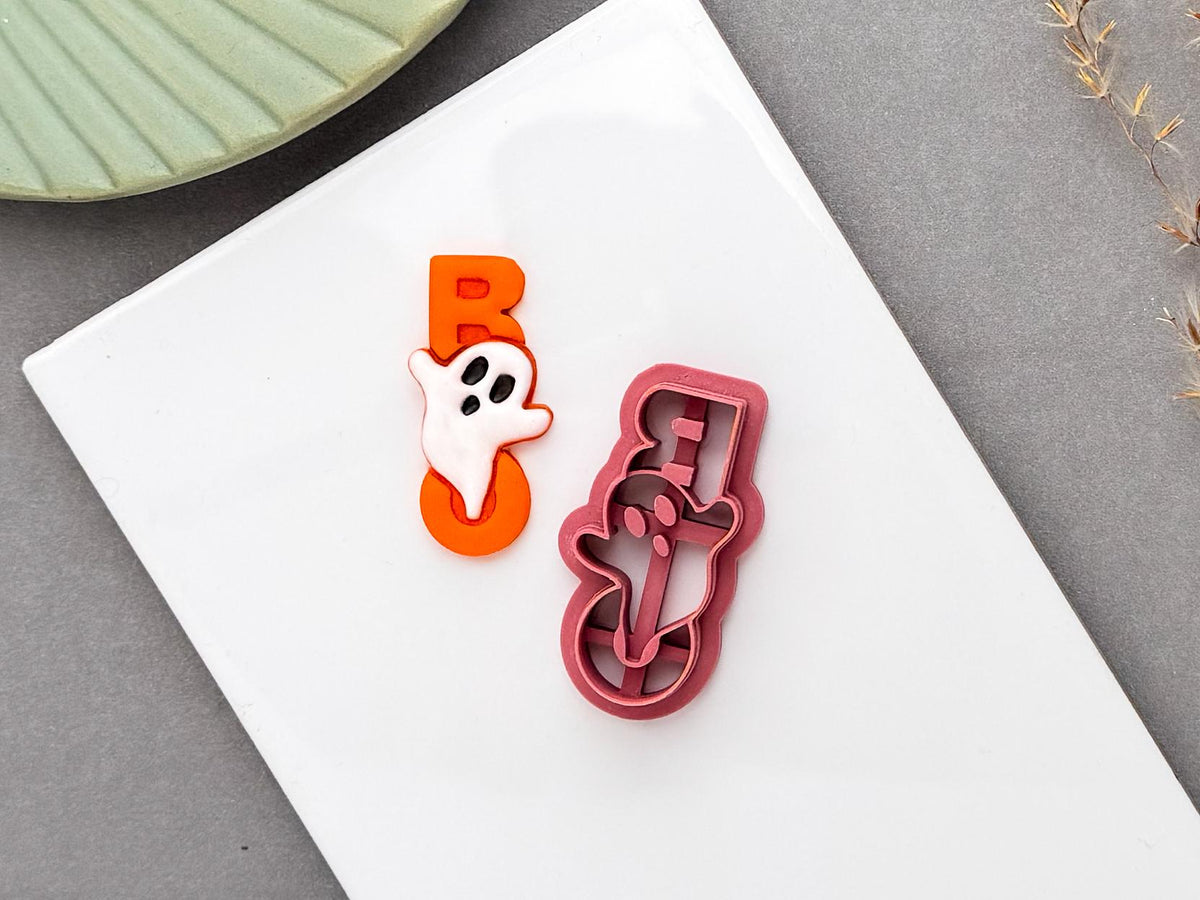 A Halloween-themed clay cutter and polymer clay ghost design, featuring a ghost decoration over a bold orange “BOO” background. The pink cutter has a detailed Halloween ghost shape with internal cutting and stamping elements, shown beside the finished clay piece on a white tile.