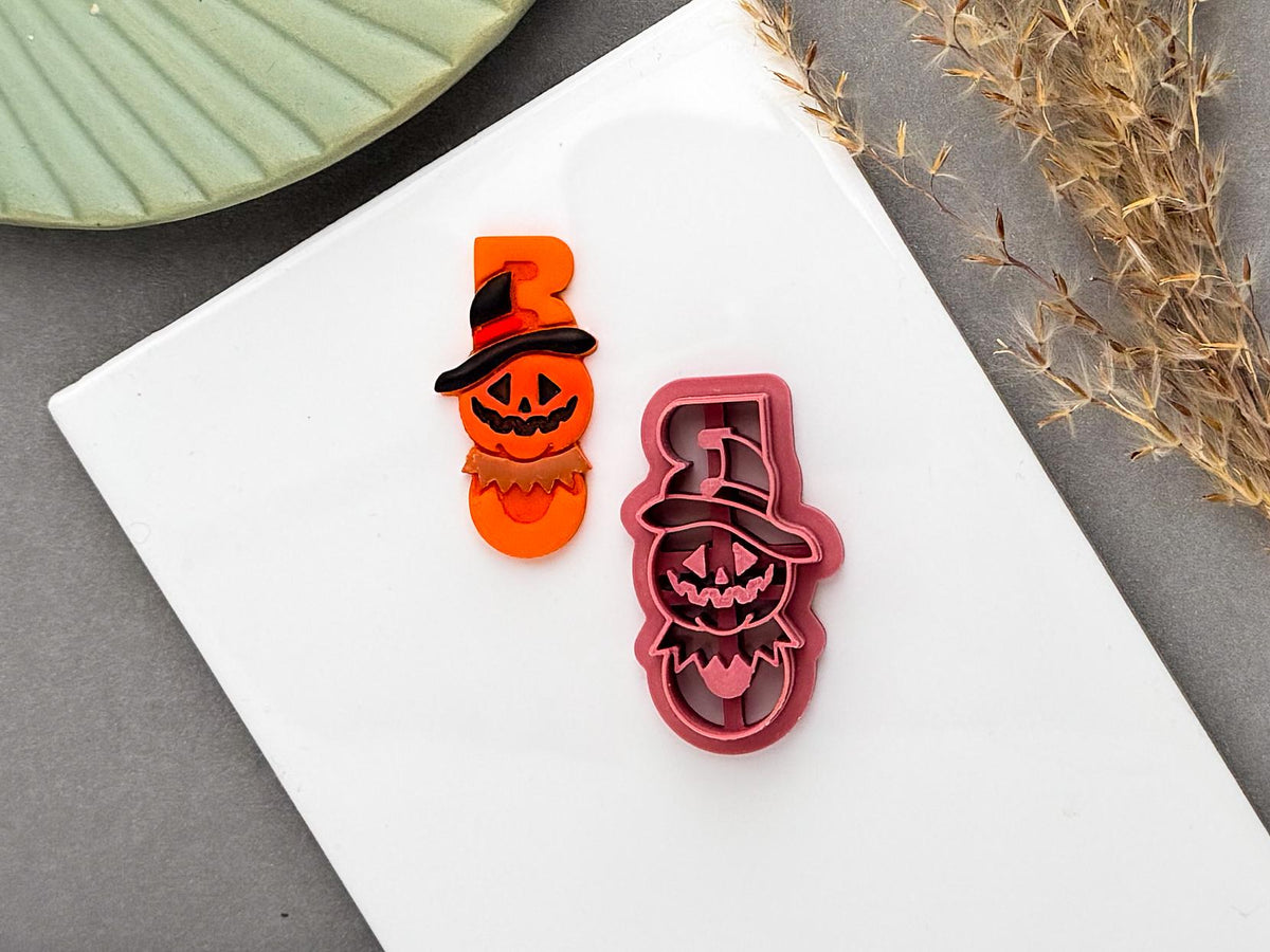 Halloween-themed clay cutter showing a spooky pumpkin character wearing a wide-brimmed witch hat, alongside the matching pink cutter on a white work surface, with autumn dried foliage in the background.