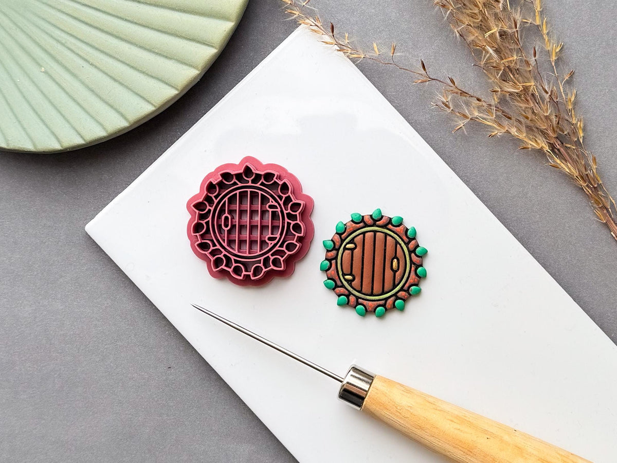 Pink 3D-printed fairy door clay cutter with a circular wooden door design and decorative stone-and-leaf border, displayed beside a finished clay fairy door on a white surface. Cottagecore, woodland, autumn and seasonal fairy garden craft theme.