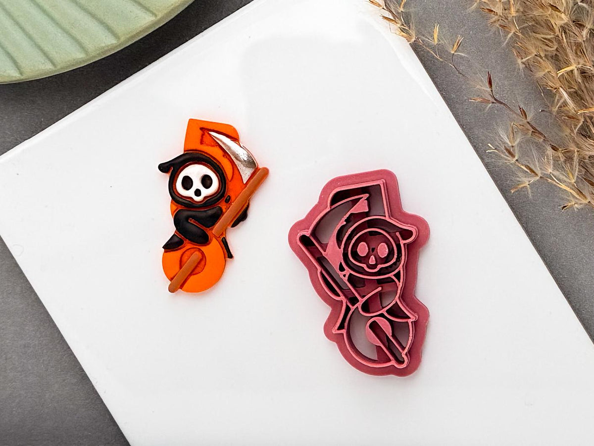 Halloween-themed clay cutter for a spooky grim reaper design, shown beside a finished orange Halloween clay piece featuring a skull-faced grim reaper with a large scythe and black hood. The pink cutter has detailed internal lines for the character and scythe, displayed on a white surface with autumn foliage in the background.