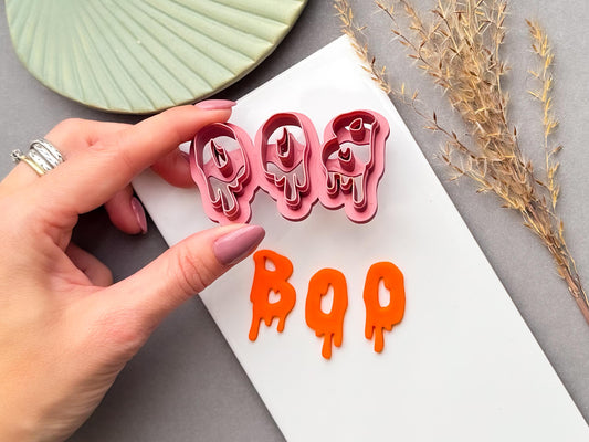 Halloween clay cutter featuring the letters “BOO” in a dripping slime-style design, held in a hand above finished orange polymer clay “BOO” letters. Spooky Halloween-inspired design for alternative jewellery and seasonal craft projects.