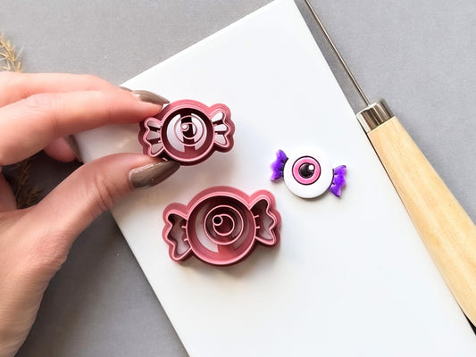 Eyeball-shaped candy clay cutters displayed in two sizes, featuring a central eyeball design within a wrapped candy shape, shown beside a finished white polymer clay eyeball candy with purple twisted ends. Halloween-inspired design displayed on a white surface.