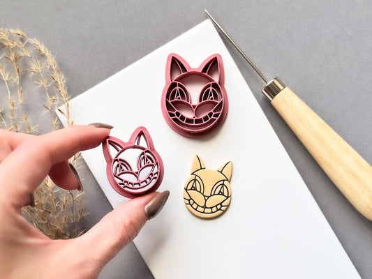 Stained glass style cat face polymer clay cutter held in a hand, with a second cutter in a larger size and a finished cream polymer clay cat face cut-out.