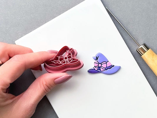 Witch’s hat clay cutter held in a hand, featuring a large bow and decorative ribbon details, shown beside a finished purple polymer clay witch’s hat with a pink bow. Spooky, witchy and Halloween-inspired design.