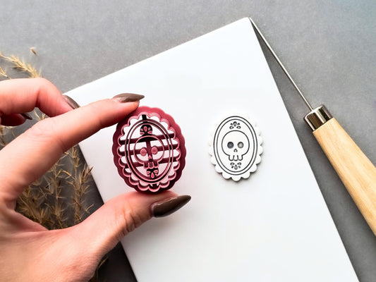 Gothic skull cameo polymer clay cutter held between fingers beside a matching finished clay cut-out, showcasing the detailed skull with floral accents, ornate oval frame and scalloped edge.