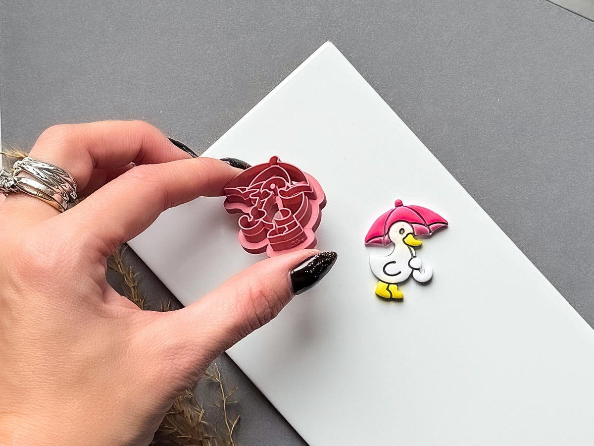 Umbrella character polymer clay cutter held between fingers to showcase the detailed umbrella and character stamp design, with a matching finished clay embellishment displayed on a white tile.