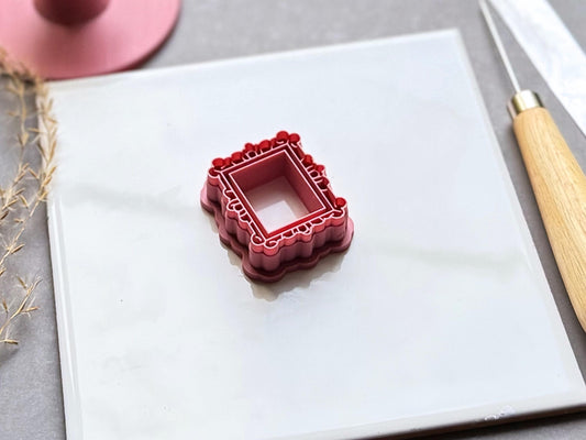 Vintage-style rectangular frame polymer clay cutter featuring an ornate scalloped border with decorative loop and bead details, displayed on a white tile.