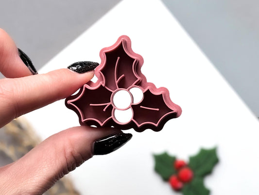 Christmas holly polymer clay cutter held between fingers to showcase the detailed three-leaf design, central berry cluster and fine leaf vein stamp lines, with the matching finished clay cut-out visible in the background.