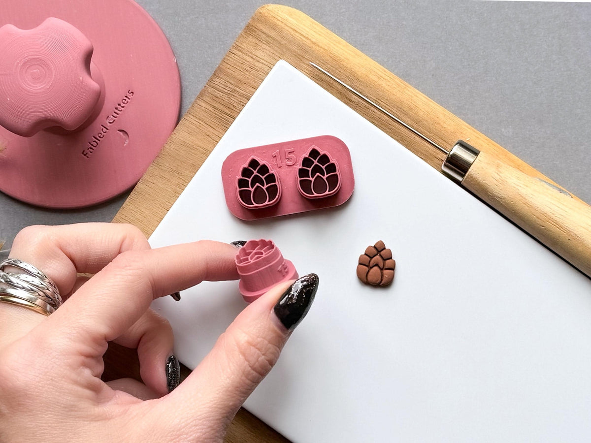 User attachment
Pine cone polymer clay stamp cutter set with two matching pine cone designs on a joined holder, shown alongside a hand-held single cutter and a finished clay pine cone embellishment. The image highlights the detailed scale pattern and internal size marking.
