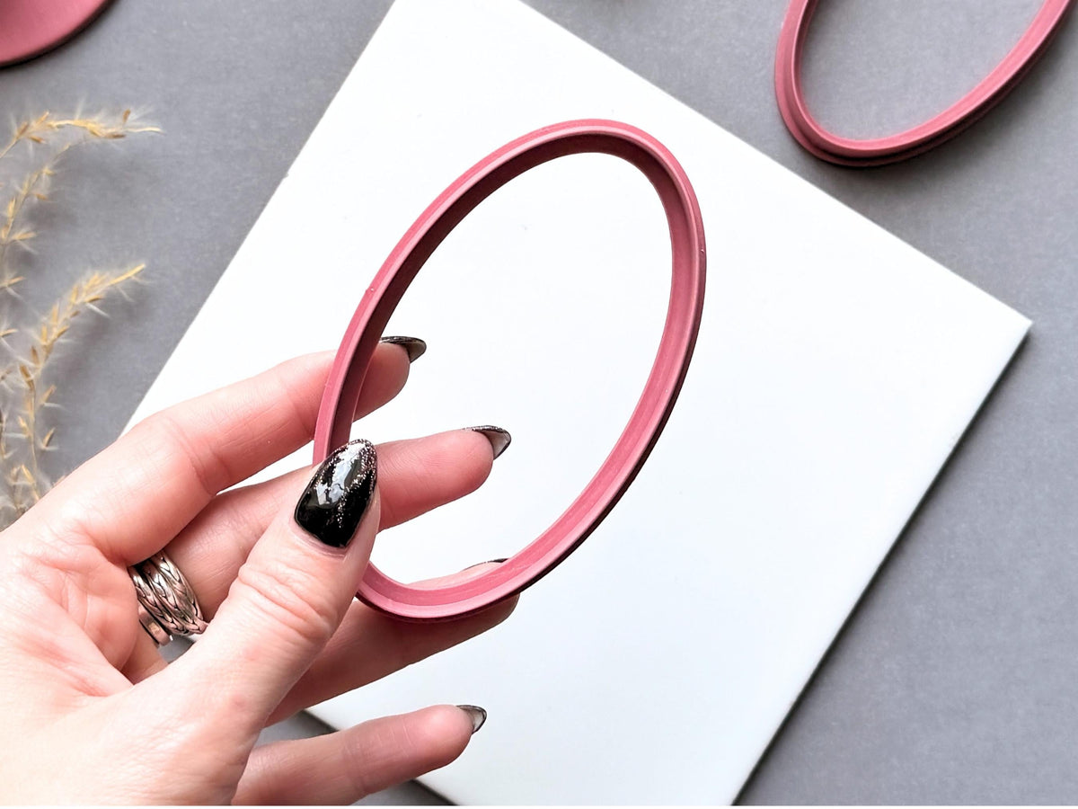 Large oval polymer clay cutter held between fingers to showcase its elongated shape, smooth inner outline and raised cutting wall, with another oval cutter visible in the background.