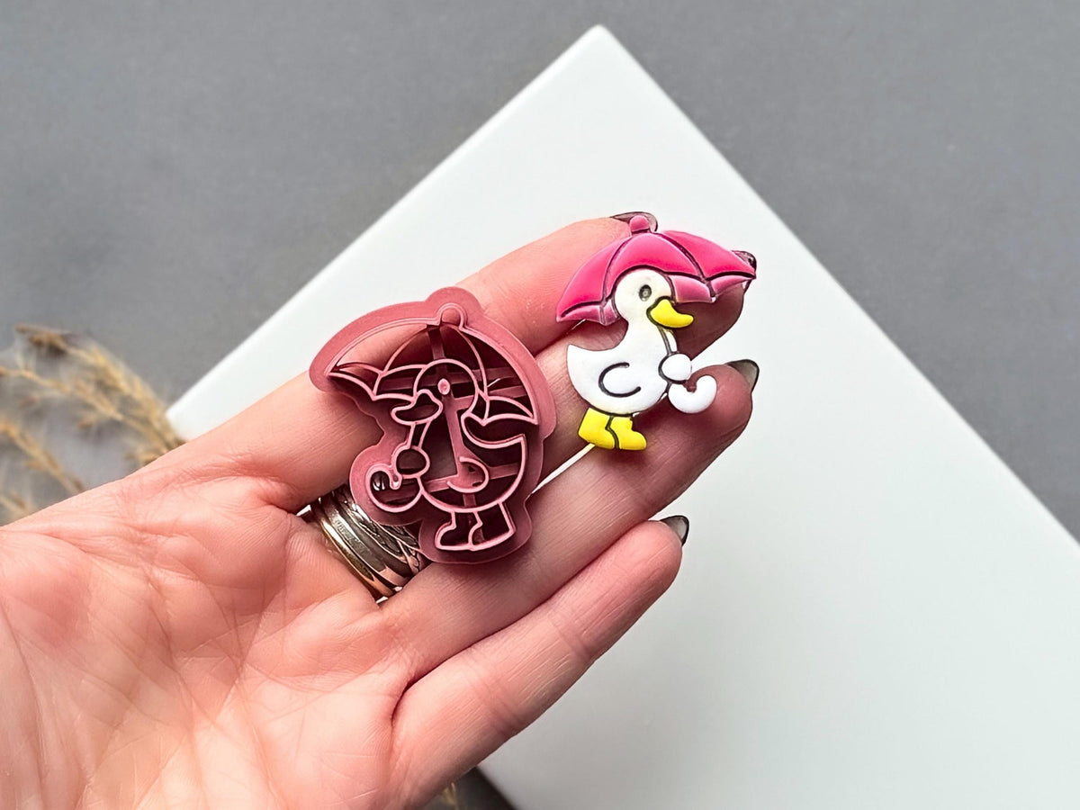 polymer clay cutter in the shape of a duck holding an umbrella, this cutter is for making polymer clay jewellery and accessories. The cutter is shown in a hand for scale along side a finished clay cut out earrinng.