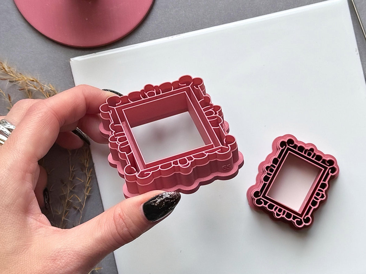 Vintage-style rectangular frame polymer clay cutters in two sizes, with ornate scalloped borders, decorative loops and rounded corner details, held by hand to showcase the larger cutter and its internal size marking.