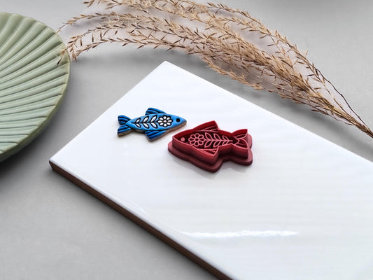 Side view of a blue folk art fish clay charm beside a dusty pink ornamental fish polymer clay cutter on a white ceramic tile.