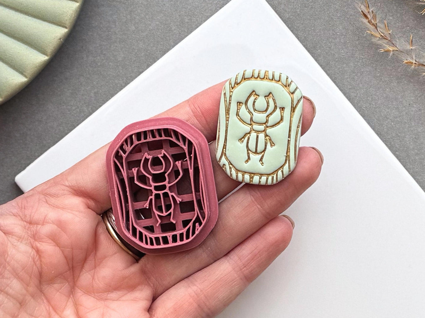 Scarab beetle polymer clay cutter and matching finished polymer clay embellishment held in a hand for scale, featuring a detailed scarab beetle within a decorative Egyptian-inspired border.