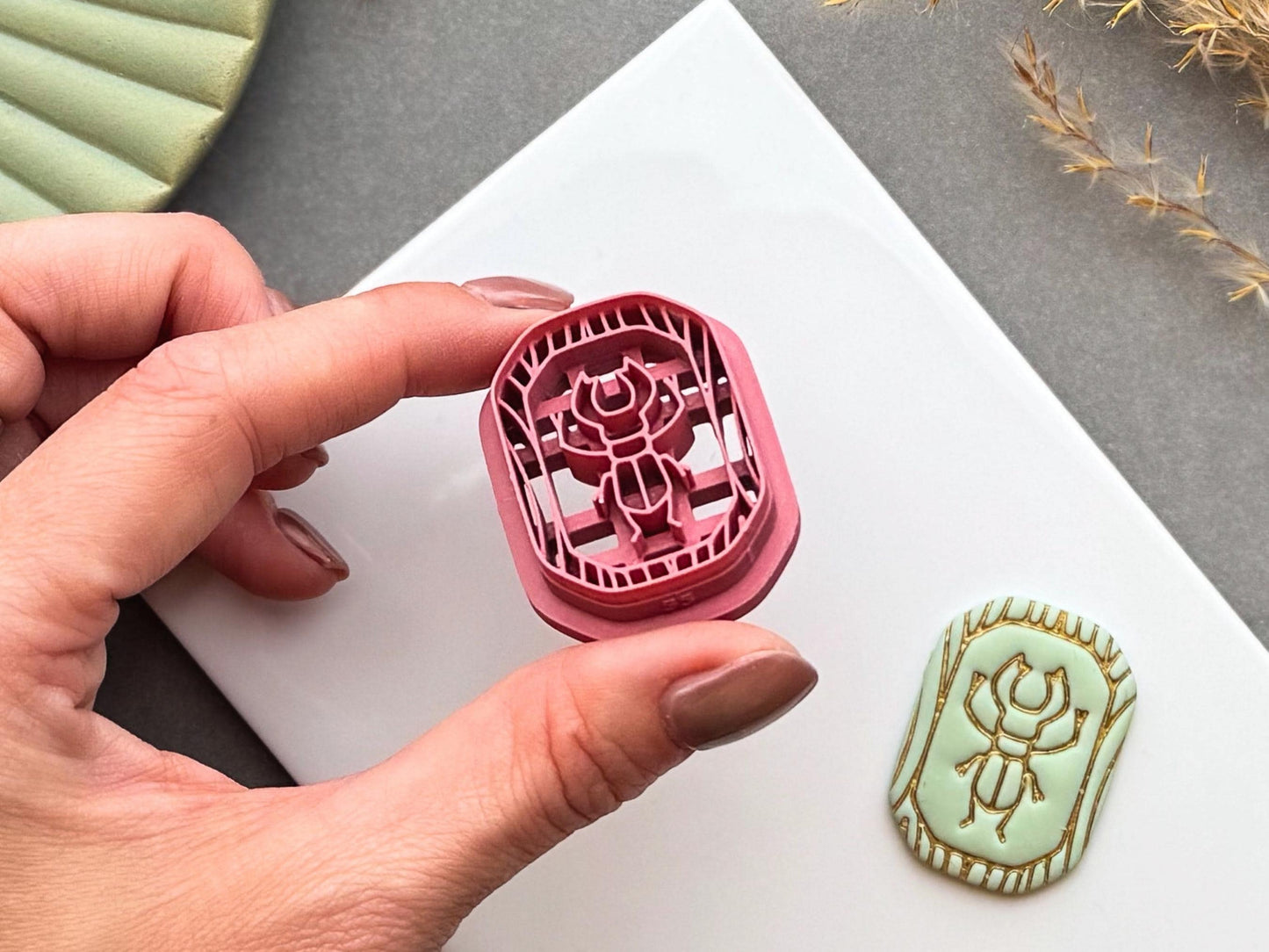 Scarab beetle polymer clay cutter held between fingers to showcase the detailed scarab design, fine cutting edge and deep stamp details, with the matching finished clay embellishment in the background.