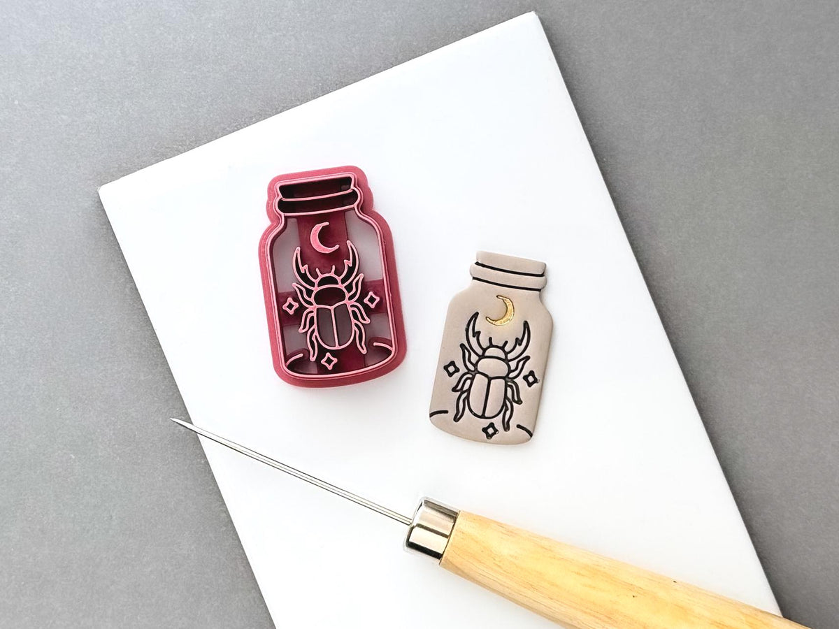 Beetle clay cutter shaped like a specimen jar, featuring a crescent moon, detailed scarab beetle and small decorative star details, shown beside a finished cream polymer clay cut-out. Gothic and celestial-inspired design.