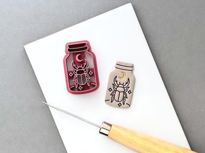 Beetle clay cutter shaped like a specimen jar, featuring a crescent moon, detailed scarab beetle and small decorative star details, shown beside a finished cream polymer clay cut-out. Gothic and celestial-inspired design.