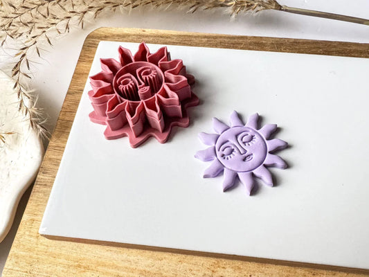 Tilted pink celestial sun face polymer clay cutter displayed with a matching lavender clay sun embellishment on a white ceramic tile.