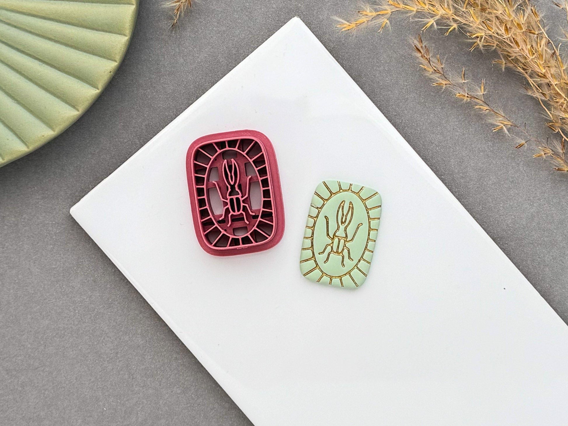 Beetle polymer clay cutter displayed beside a matching finished polymer clay embellishment featuring a simple beetle motif with an Art Deco-inspired geometric border.