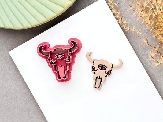 3 eyed mystical bull skull clay cutter with curved horns and detailed facial features, shown beside a finished polymer clay bull skull with painted eyes and tongue. Alternative, gothic and western-inspired design for polymer clay jewellery and craft projects.