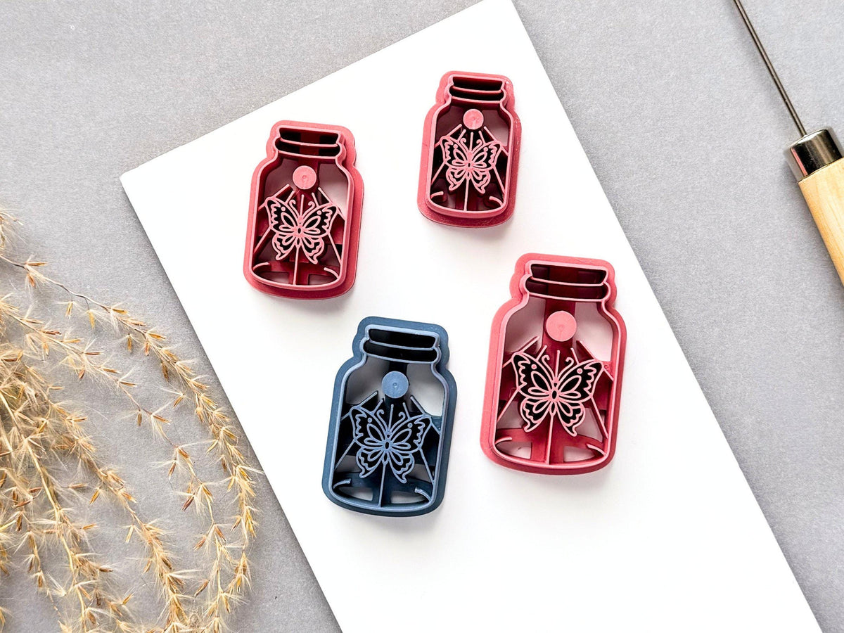 Four butterfly specimen jar clay cutters displayed in different sizes and colours, featuring detailed symmetrical butterfly designs with decorative floral details. Three pink cutters and one blue cutter are arranged on a white surface, showcasing the different available sizes.