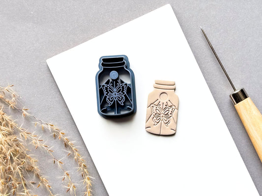 Butterfly specimen jar clay cutter featuring a detailed symmetrical butterfly design with decorative floral and celestial-inspired details, shown beside a finished cream polymer clay butterfly cut-out. Blue 3D-printed cutter displayed on a white surface.